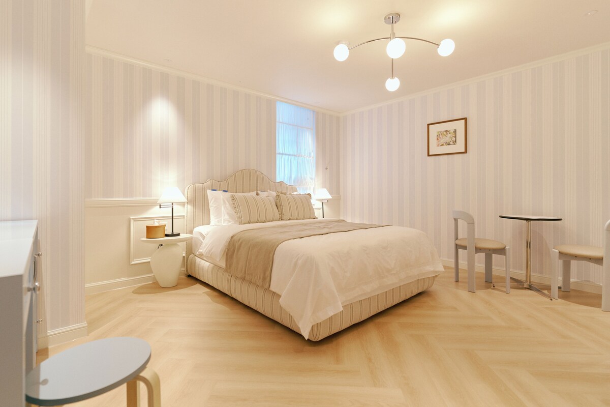 Un dormitorio con cama, mesitas de noche, una silla, una pequeña mesa y un cuadro en la pared.