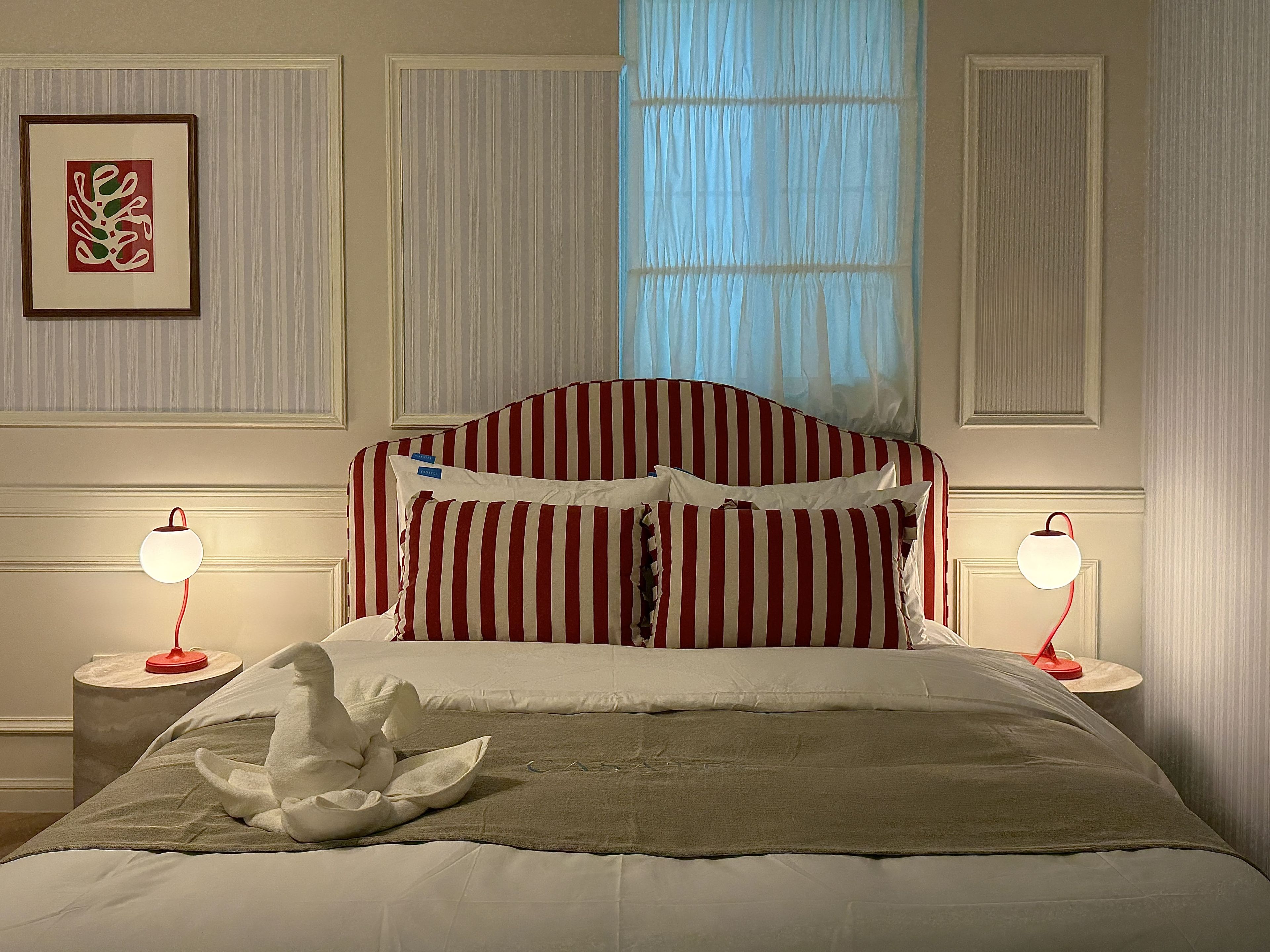 Una cama bien tendida con ropa de cama a rayas, dos lámparas de noche, un cuadro en la pared y una ventana con cortinas traslúcidas.