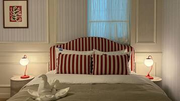 Una cama bien tendida con ropa de cama a rayas, dos lámparas de noche, un cuadro en la pared y una ventana con cortinas traslúcidas.