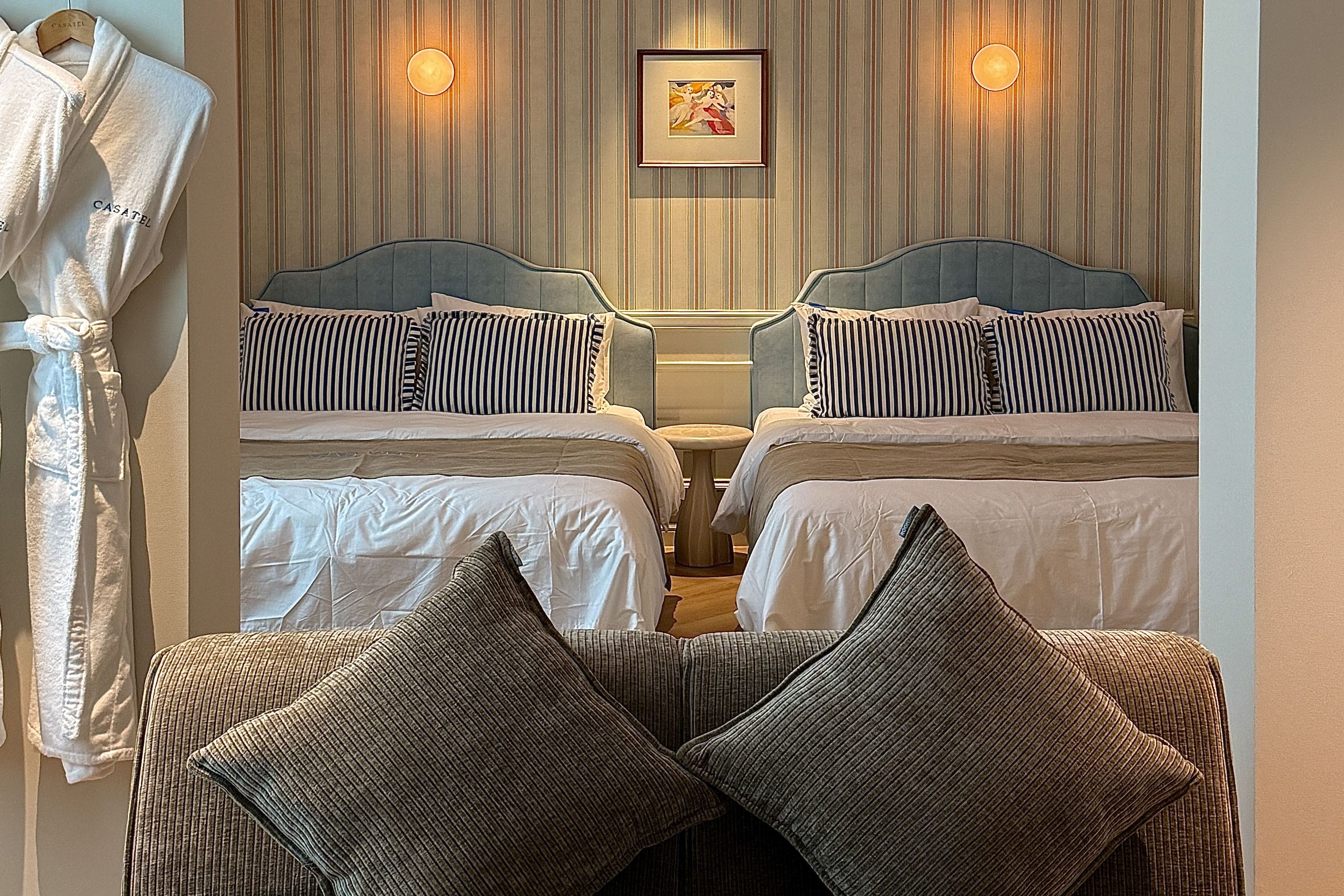 Quarto de hotel com duas camas, área de estar com uma poltrona e almofadas, roupão pendurado e quadro na parede.