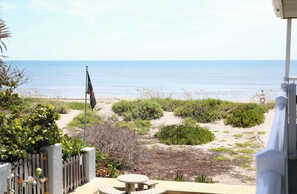 Eine Terrasse mit Tisch und Stühlen, eine Fahnenstange mit einer Flagge sowie Blick auf den Strand und das Meer.
