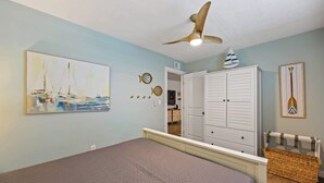 Ein Schlafzimmer mit einem Bett, einem Deckenventilator, einem Gemälde mit Segelbooten, einem Kleiderschrank und einem an der Wand befestigten Surfbrett.