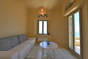 A room with a white sofa, a small round table with a blue bowl, a hanging light fixture, a window with a view, and a sliding glass door. - Dioni Tinos, sea view elegant villa (Kardiani)
