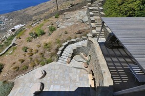 A stone retaining wall with a staircase leading down to a paved area with potted plants. - Dioni Tinos, sea view elegant villa (Kardiani)