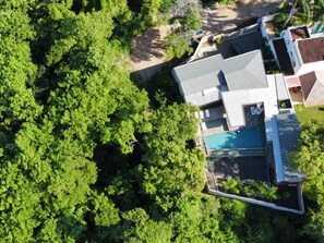 Vista aérea de una zona residencial con una casa moderna, techo inclinado, piscina y exuberantes árboles verdes rodeando la propiedad.