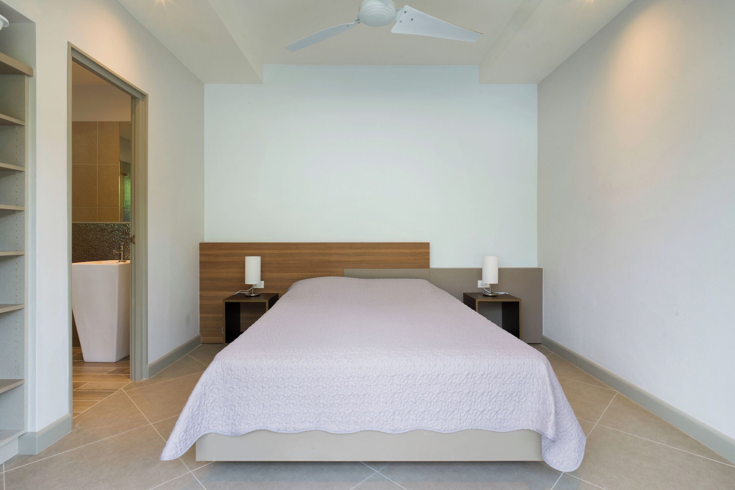 Une chambre à coucher avec un lit, des tables de chevet, un ventilateur de plafond et une salle de bain visible à travers une porte ouverte.