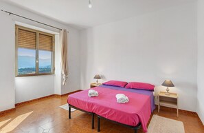 Ein Schlafzimmer mit einem Bett, zwei Nachttischlampen, einem Fenster mit Jalousien und Blick ins Freie.