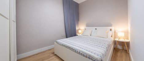 Un dormitorio con cama, lámparas de noche, una ventana con cortinas y suelo de madera.