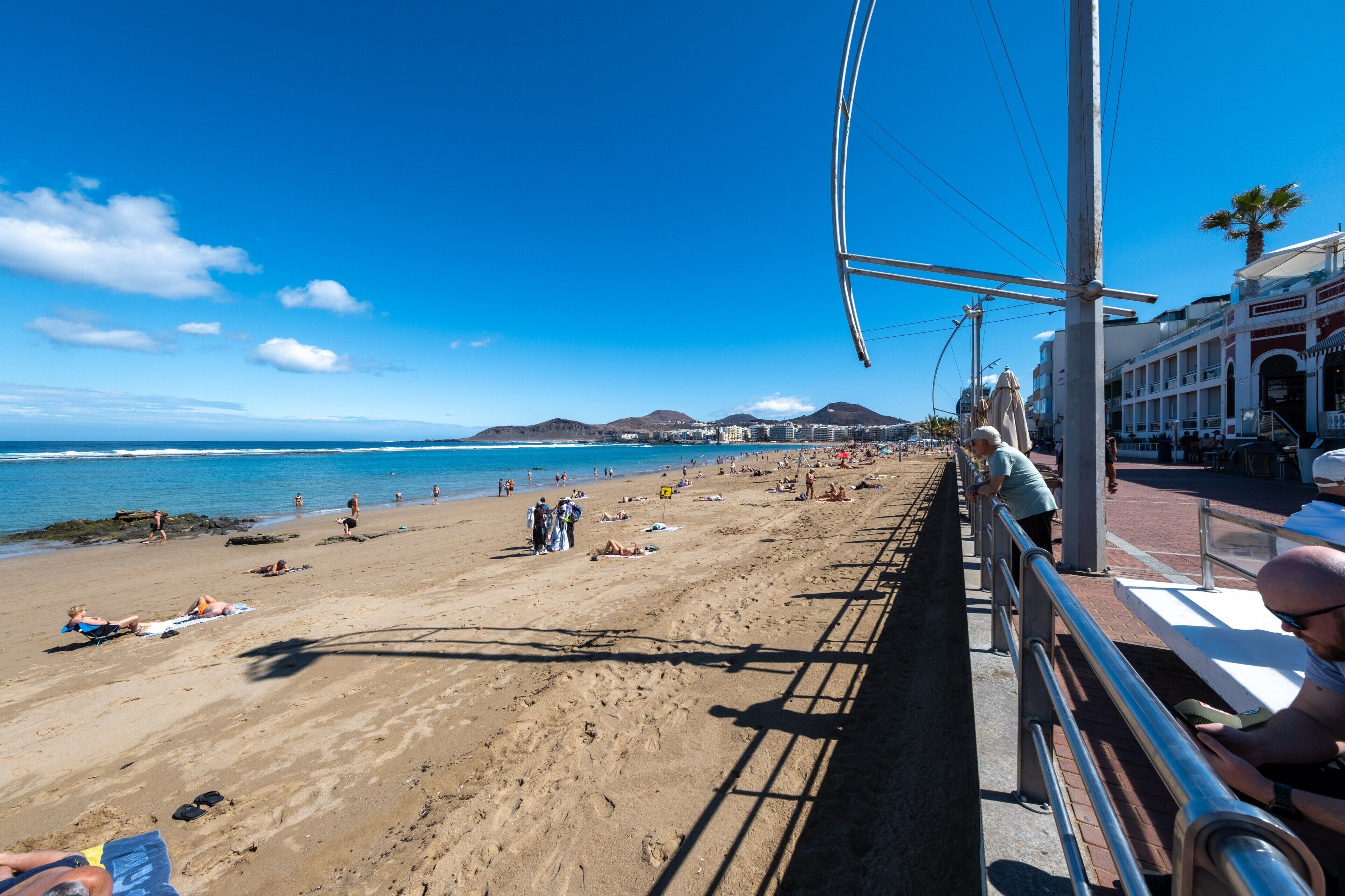 Una playa con gente tomando el sol y nadando, cielo despejado y edificios al fondo.