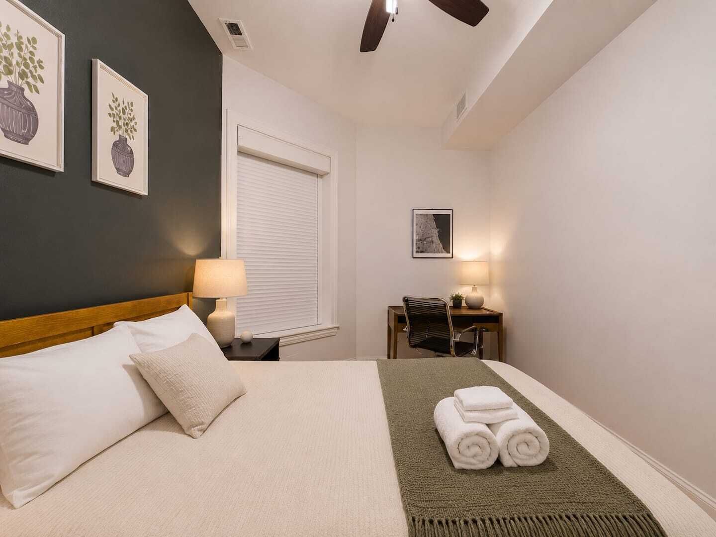A hotel room with a bed, bedside table, desk, chair, and artwork on the walls.
