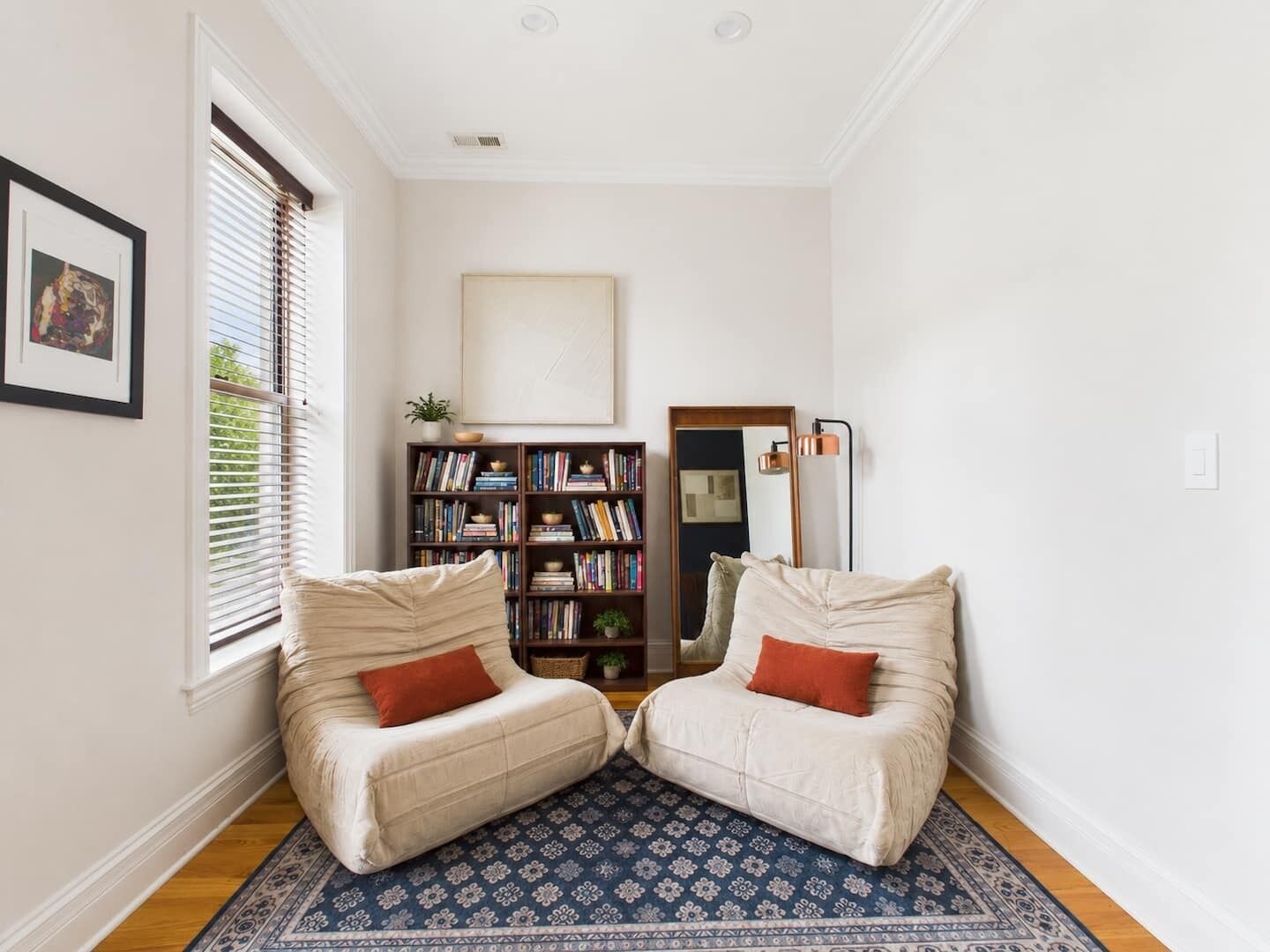 Una habitación con dos sillones color beige, una estantería, un espejo y una alfombra estampada.