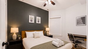 Quarto de hotel com uma cama, duas luminárias de cabeceira, uma escrivaninha com cadeira, um ventilador de teto e duas quadros na parede.