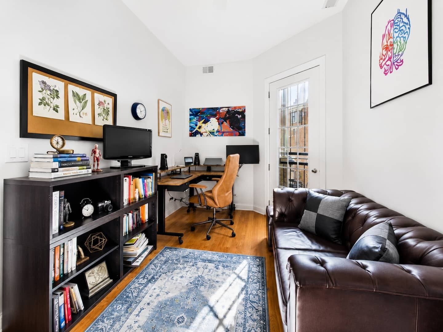A modern living room with a leather sofa, a bookshelf, a desk with a computer, and a large window.