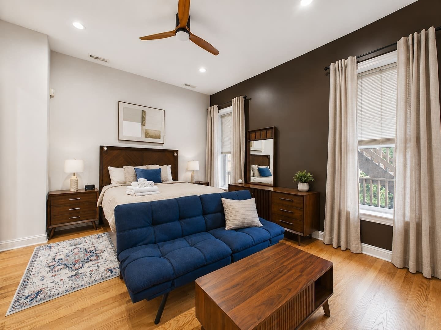 Un dormitorio con cama, mesitas de noche, un sofá azul, una mesa de centro de madera, un ventilador de techo y una ventana con cortinas.