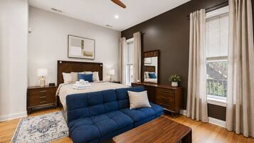 Un dormitorio con cama, mesitas de noche, un sofá azul, una mesa de centro de madera, un ventilador de techo y una ventana con cortinas.
