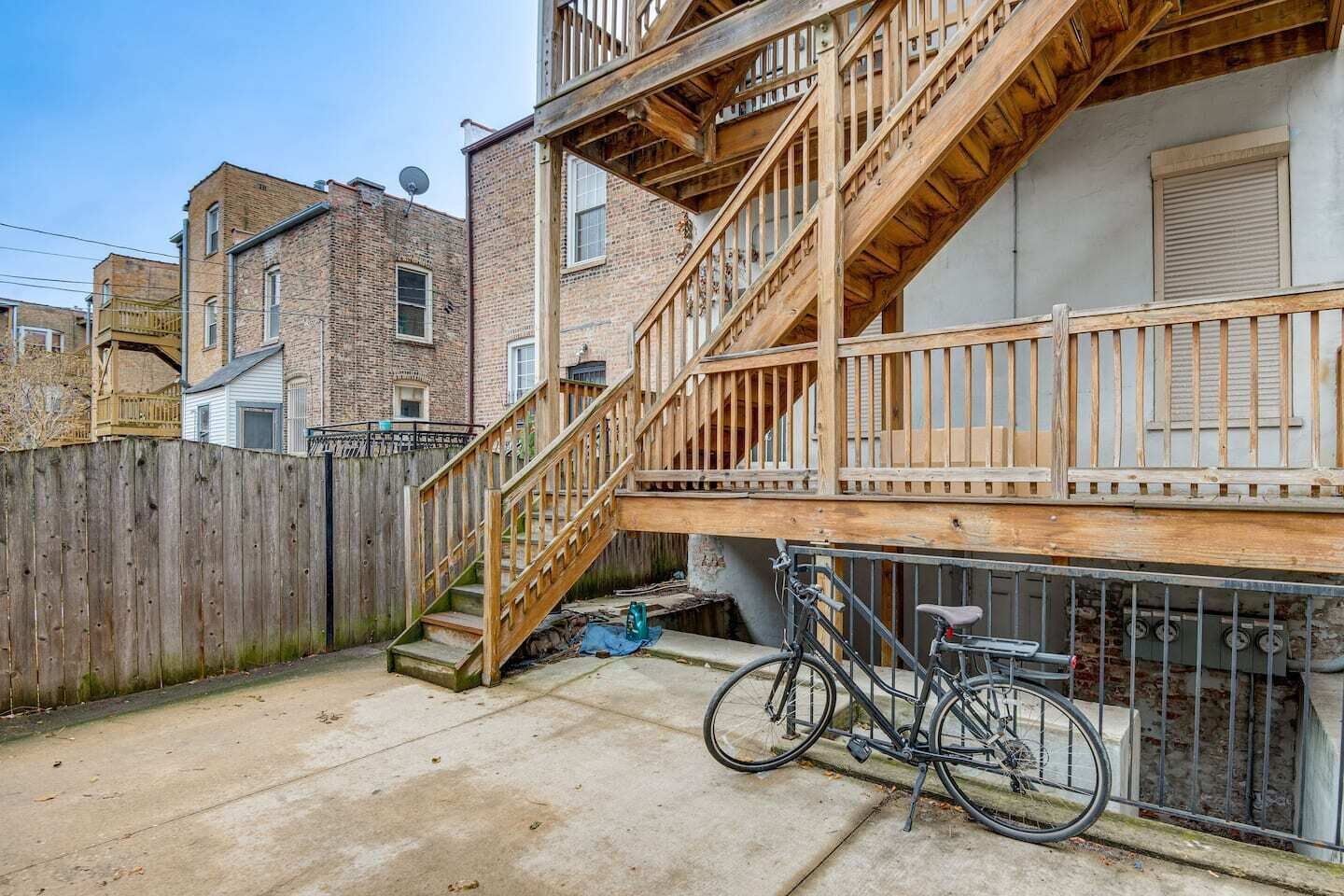 A residential building with a wooden staircase leading to a balcony, a bicycle parked outside, and a wooden fence.