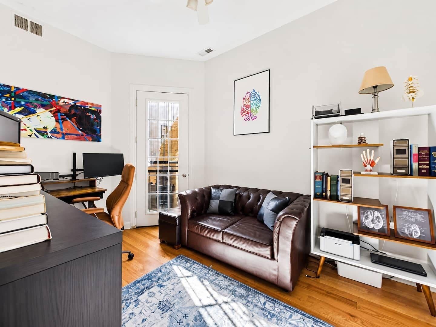 A modern living room with a leather sofa, a desk with a computer, a bookshelf, and a large abstract painting.