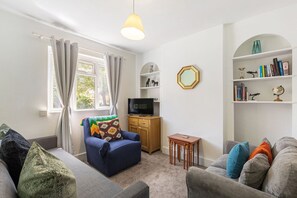 A living room with a blue sofa, a grey sofa, a wooden cabinet, a small table, and a bookshelf with various items. - GuestReady - Charming Kingston Home with Garden (London)