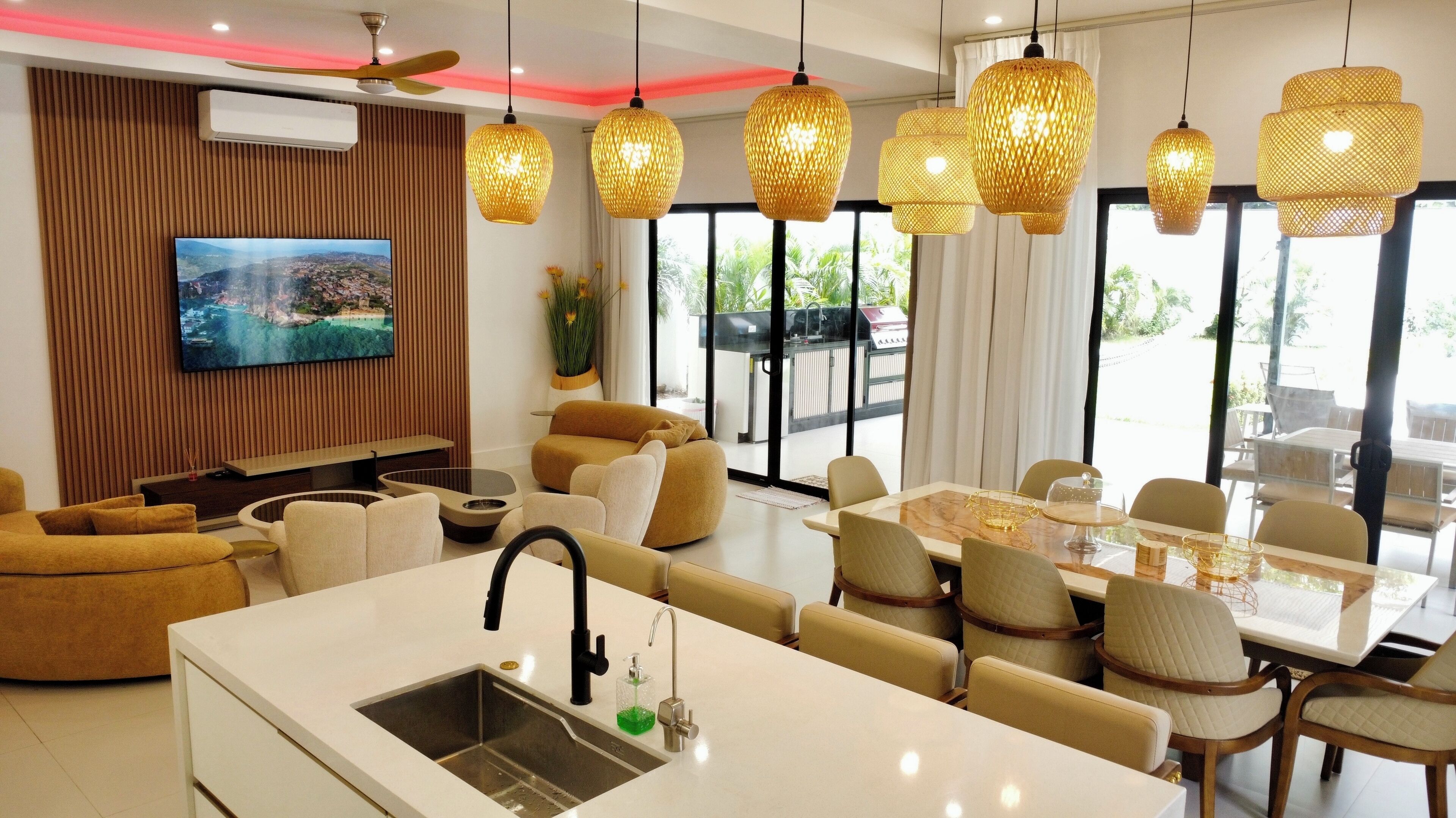 A modern hotel room with a flat-screen TV, a dining area with a table and chairs, a sofa, and a kitchen area with a sink and a bottle of soap.