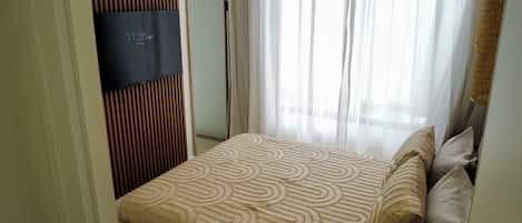 Quarto de hotel com cama, janela com cortinas leves e televisão na parede.