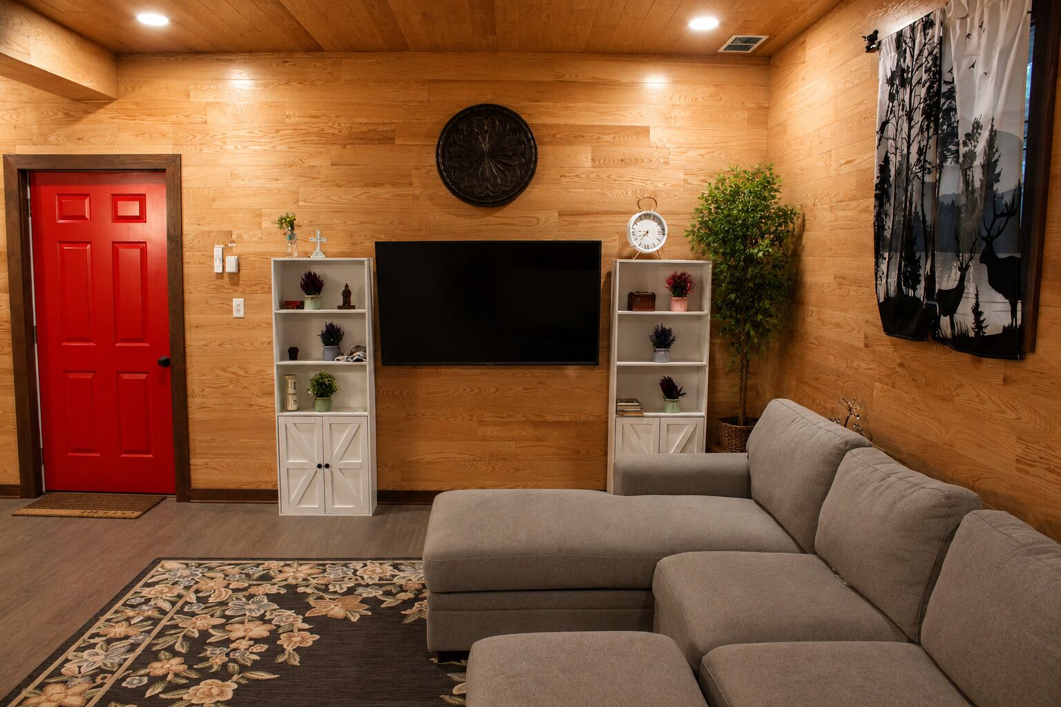 A living room with a sectional sofa, a flat-screen TV, wooden walls, a red door, and two white shelving units with plants.