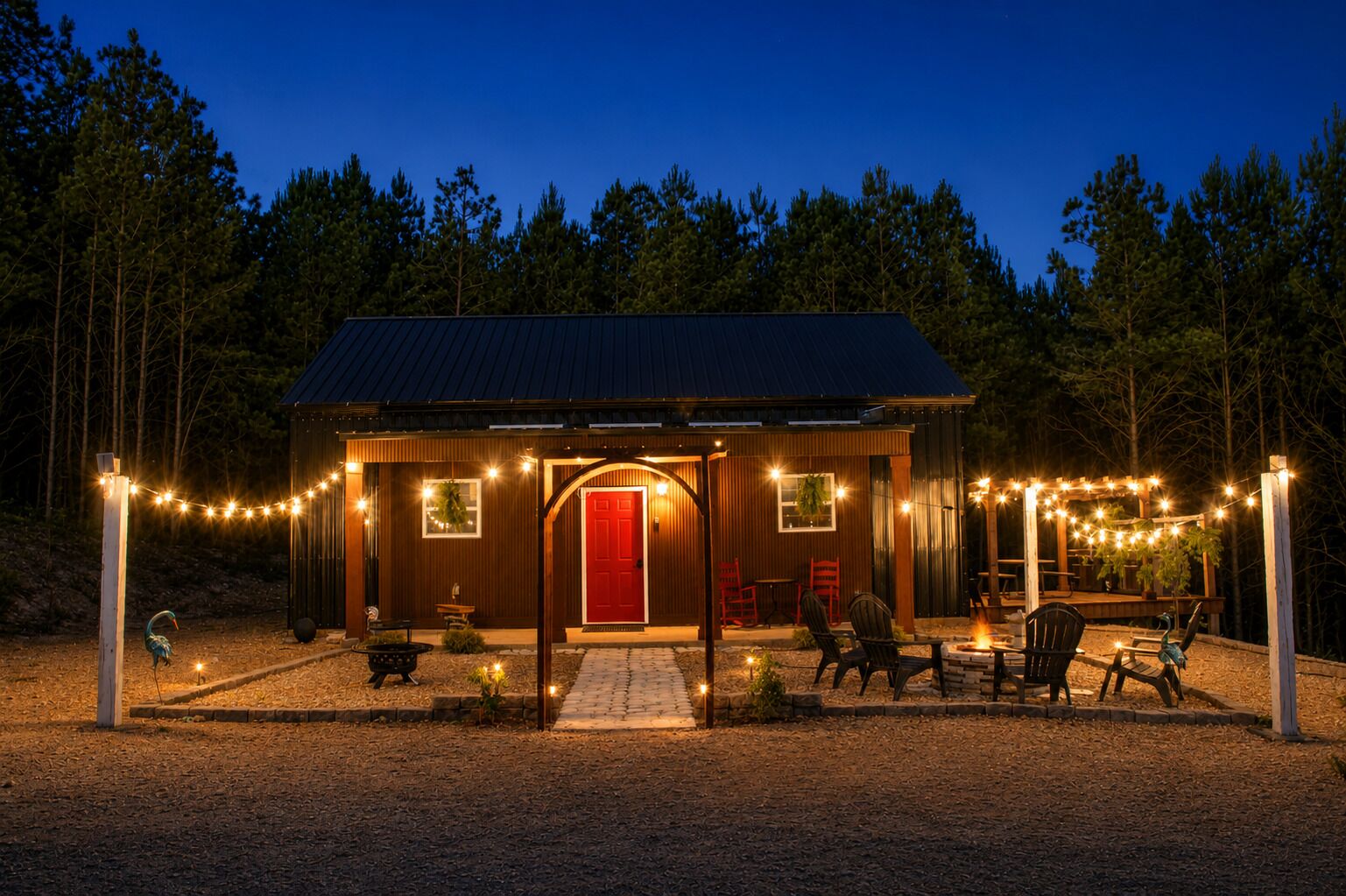 A small wooden cabin with a red door, surrounded by a deck with outdoor seating and string lights.