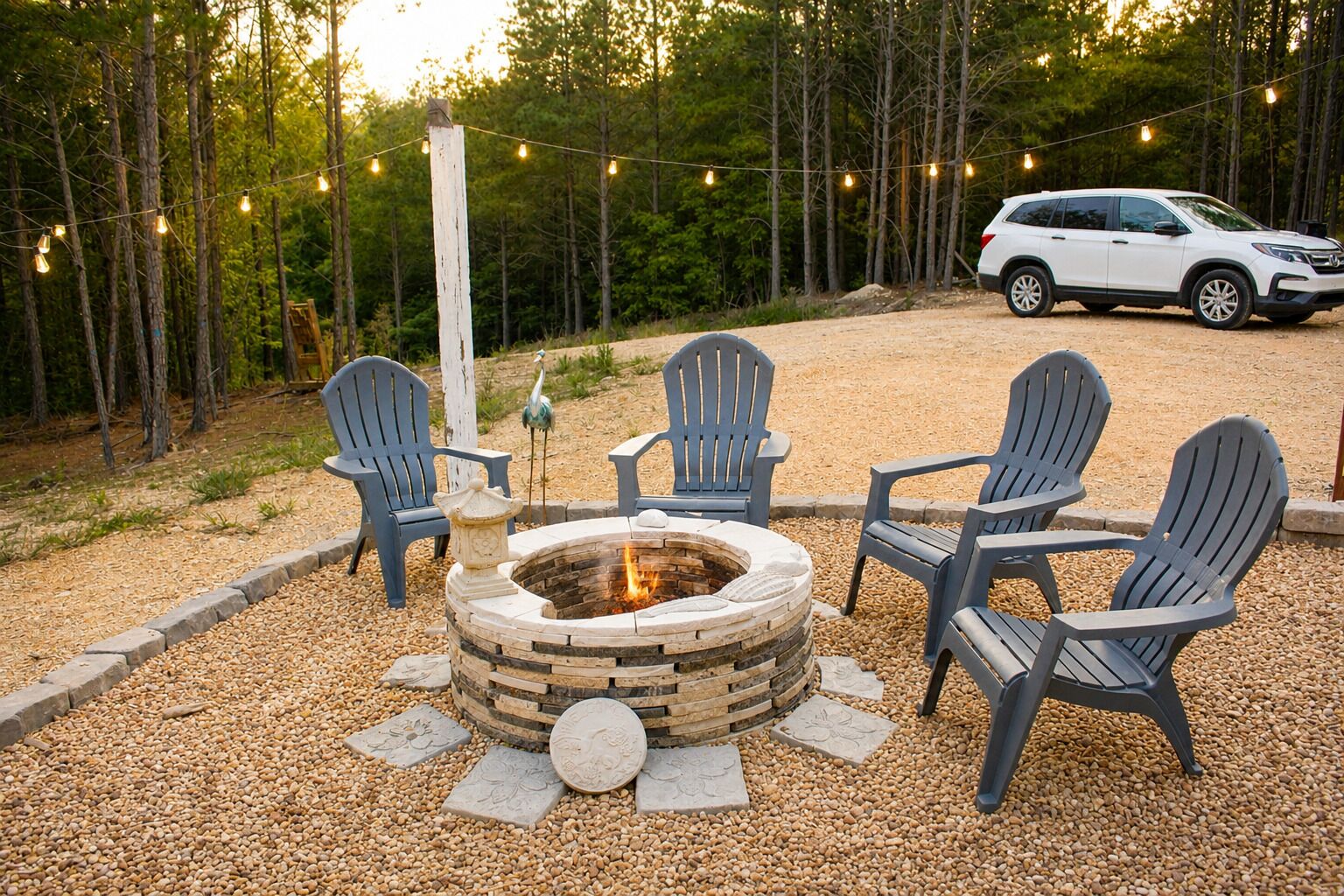 A fire pit with seating, surrounded by pebbles, with a vehicle in the background and trees.
