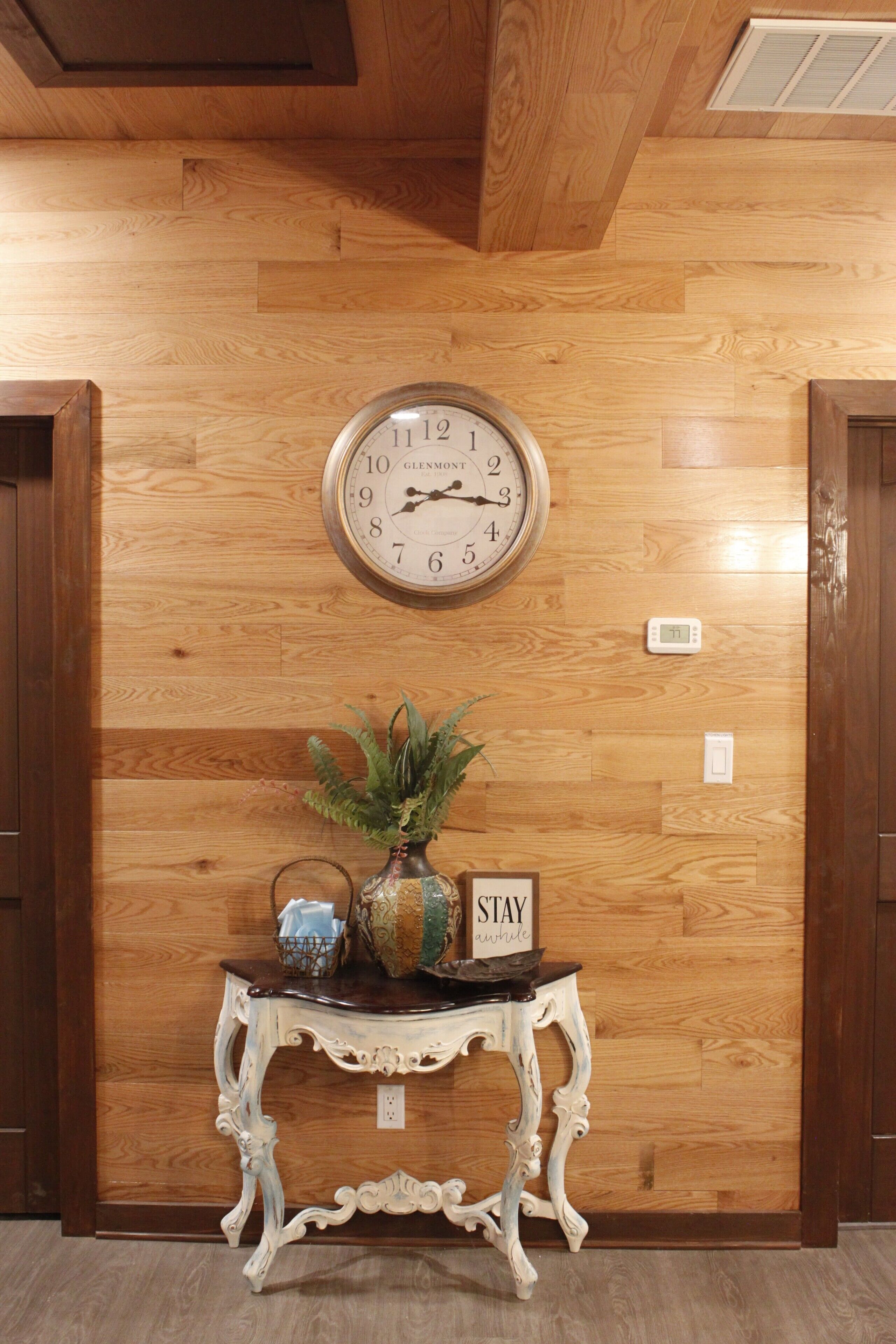 A wooden wall with a clock, a small table with a plant, and a sign that says 'STAY awhile'.
