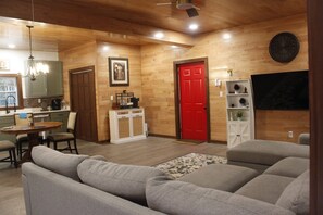 A living room with a sofa, a dining area with a table and chairs, a kitchen with cabinets, and a red door.