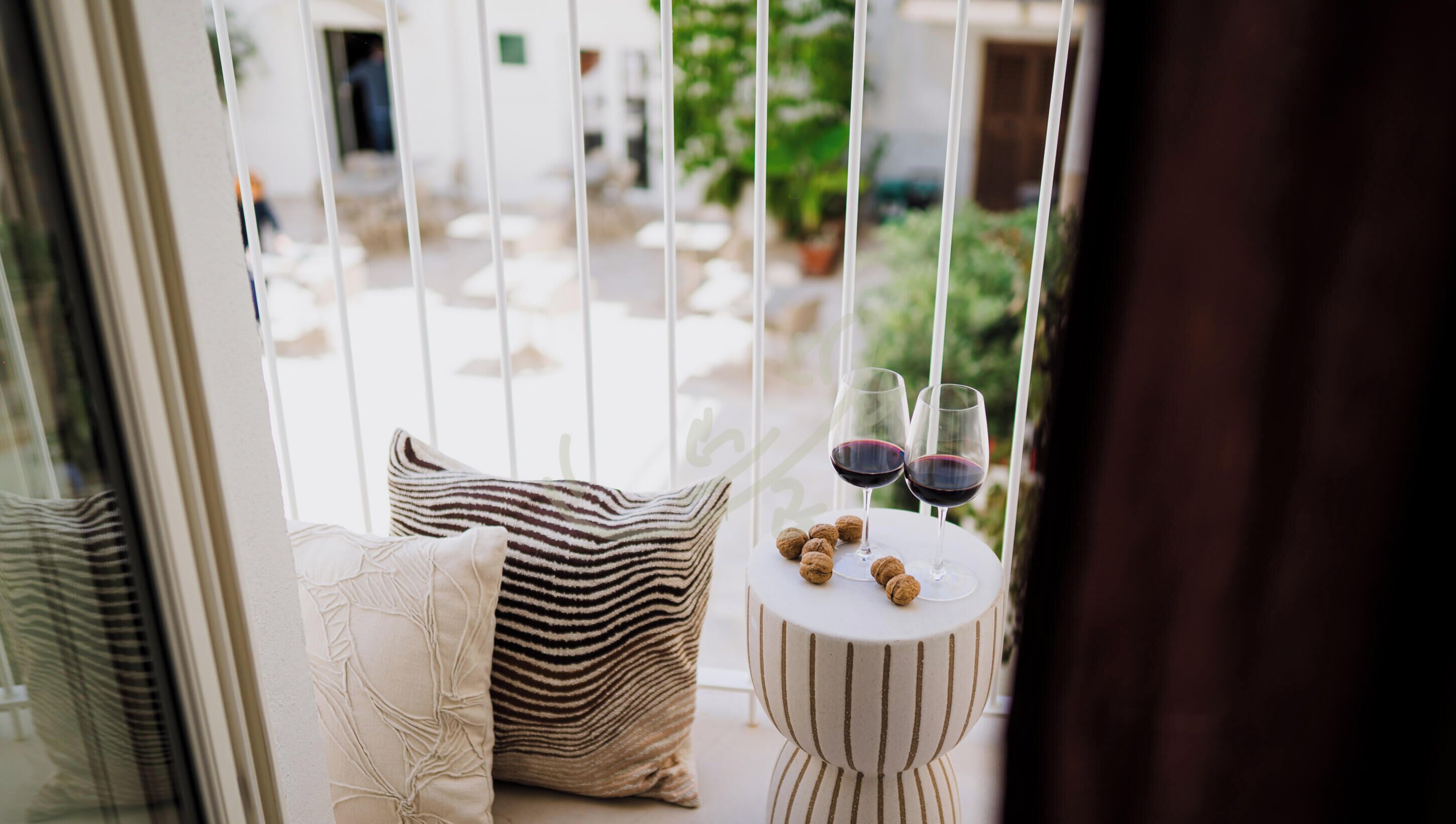 A balcony with a striped cushion, a small table with two wine glasses and walnuts, and a view of a courtyard.