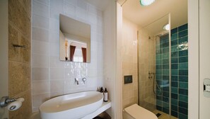 Kylpyhuoneessa on valkoinen pesuallas, peili, sinisillä laatoilla päällystetty suihku ja wc-istuin.