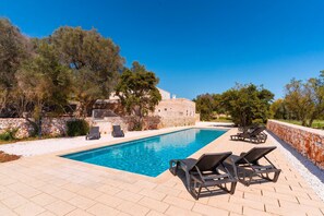A swimming pool with lounge chairs, a stone wall, and a building in the background. - Masseria Grotta Antica 6+2, Emma Villas (Ostuni)
