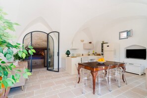 Ein Essbereich mit Holztisch und transparenten Stühlen, eine Küche mit weißen Schränken und Geräten sowie ein an der Wand befestigter Fernseher.