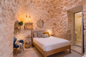 A bedroom with a stone wall, a bed with a wooden frame, a bedside table with a lamp, a chair, and a small plant. - Masseria Grotta Antica 6+2, Emma Villas (Ostuni)