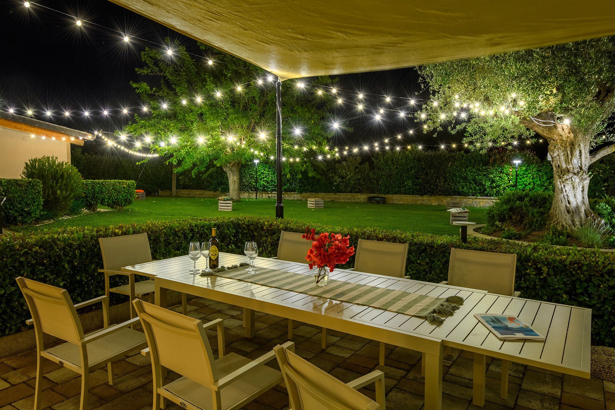 Ein Essbereich im Freien mit Tisch und Stühlen, Lichterketten über dem Kopf und einem gepflegten Garten im Hintergrund.