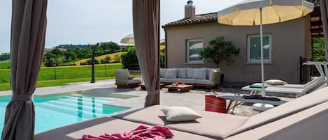 Zona junto a la piscina con sofá y sillones de descanso, una mesa con una bebida y vista a una casa y el paisaje.