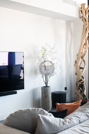 Ein modernes Wohnzimmer mit einem wandmontierten Flachbildfernseher, einer hohen Vase mit weißen Blumen, einem schwarzen Lautsprecher und einer einzigartigen Holzskulptur.
