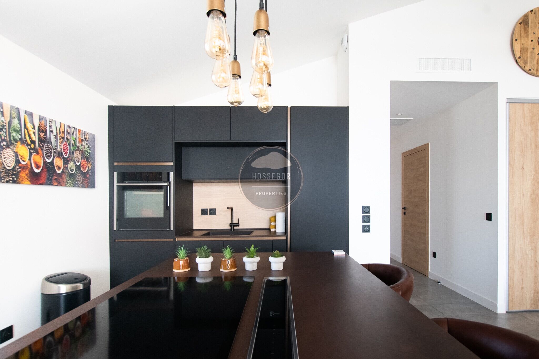 A modern kitchen with a central island, built-in oven, and hanging light fixtures.