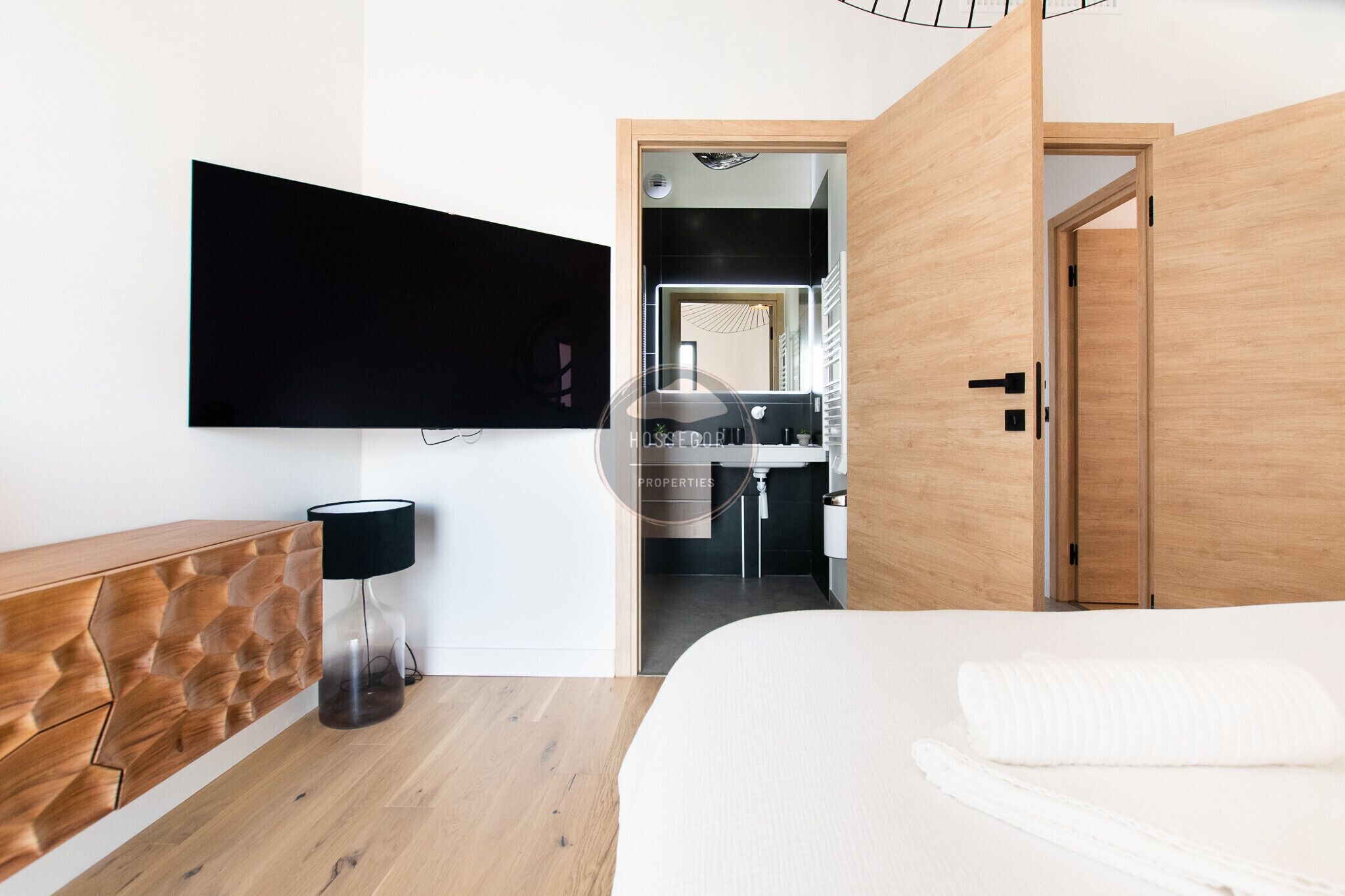Habitación de hotel moderna con televisor de pantalla plana montado en la pared, cómoda de madera, lámpara de noche y cama grande.