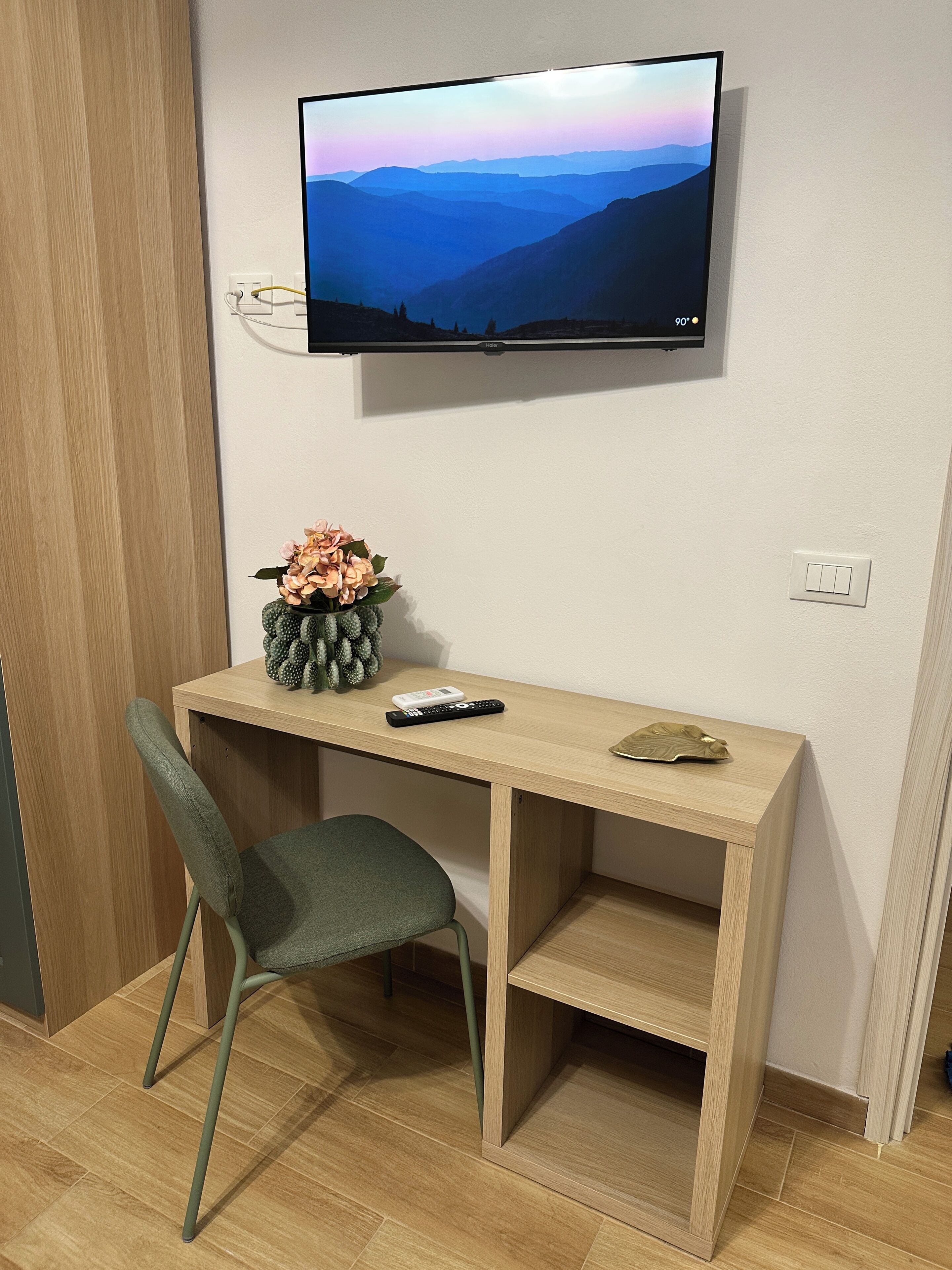 A room with a wooden desk, a chair, a television mounted on the wall, and a small decorative plant.
