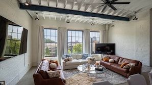 A spacious living room with a high ceiling, large windows, a flat-screen TV, and leather sofas.