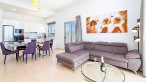 Een moderne woonkamer met een grijze hoekbank, een glazen salontafel met een fles, en een eethoek met paarse stoelen.