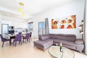 Ein modernes Wohnzimmer mit einem grauen Ecksofa, einem Glastisch mit einer Flasche und einem Essbereich mit violetten Stühlen.