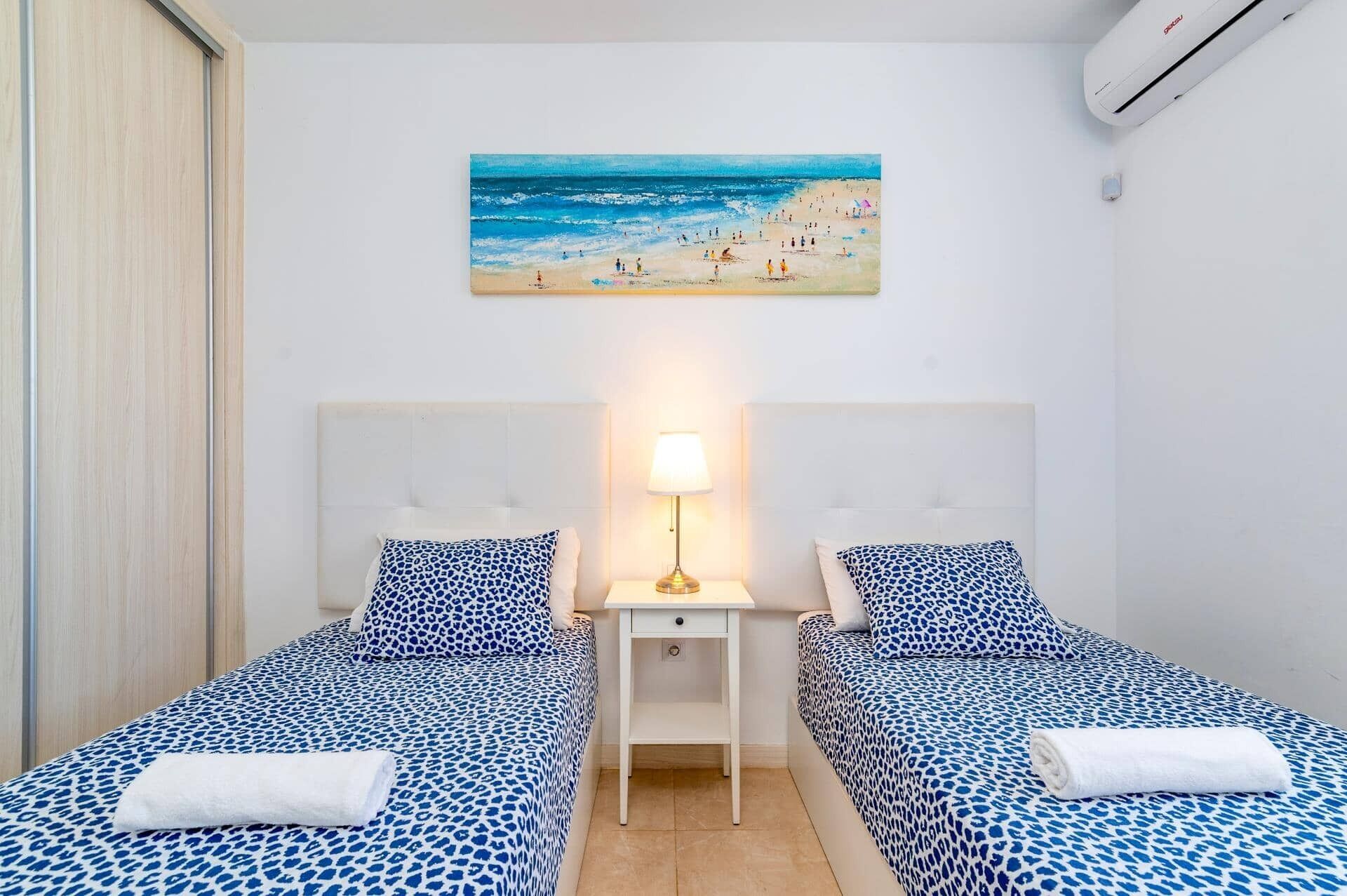 Habitación de hotel con dos camas, una mesita de noche, una lámpara, un cuadro con una playa y un equipo de aire acondicionado.