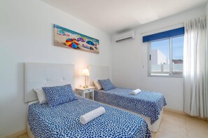 Ein Hotelzimmer mit zwei Betten, einem Nachttisch, einer Lampe, einem Gemälde, einer Klimaanlage und einem Fenster mit Vorhängen.