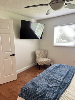 Ein Schlafzimmer mit einem Bett, einem Sessel, einem an der Wand befestigten Fernseher und einem Deckenventilator.
