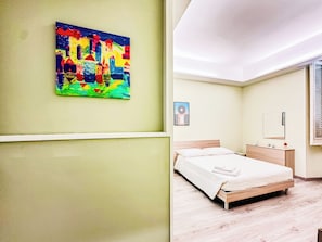 Un dormitorio con cama, mesita de noche, espejo y un cuadro de colores en la pared.