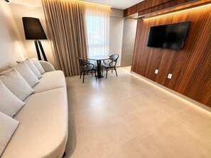 Una habitación de hotel moderna con sofá, una mesa de comedor con sillas, un televisor de pantalla plana montado en una pared de madera y una ventana con cortinas.