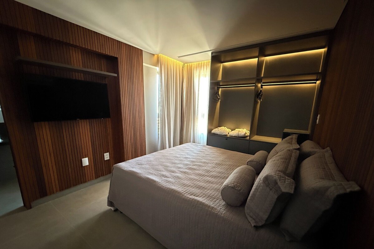 Een moderne slaapkamer met een groot bed, een flatscreen-tv aan de muur en een inbouw hoofdeinde met geïntegreerde verlichting.