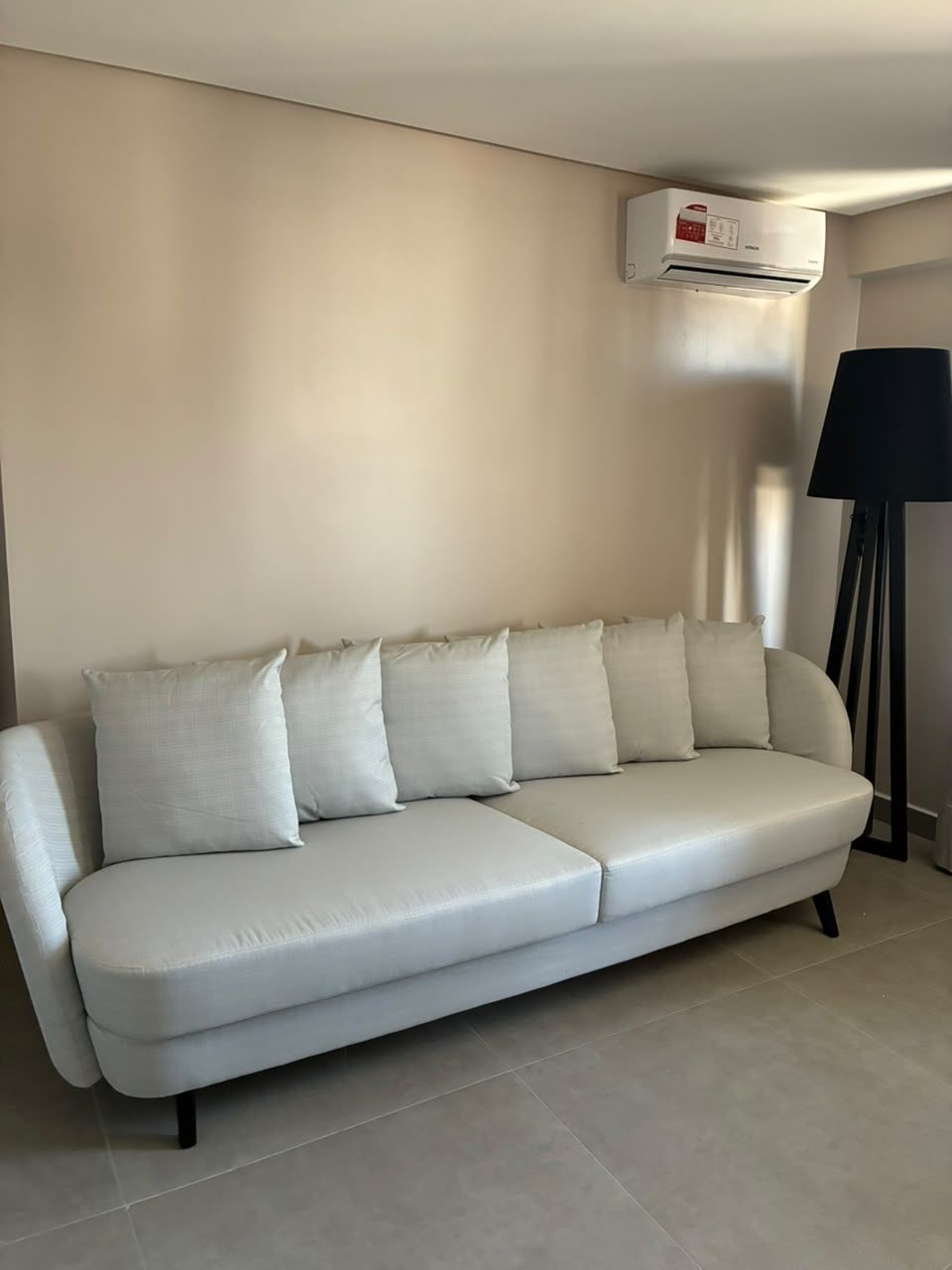 Um sofá claro com várias almofadas em frente a uma parede lisa, uma luminária de pé e um ar-condicionado instalado no teto.